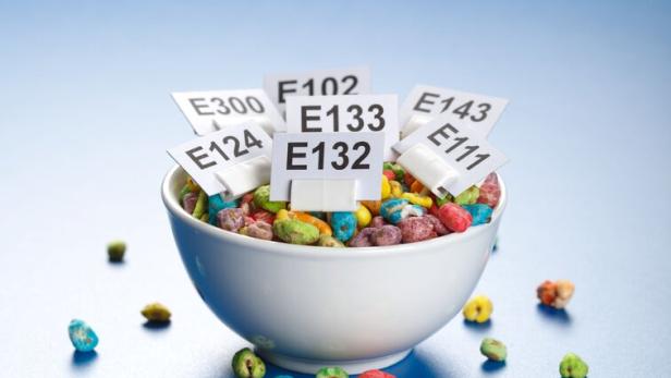 Eine Schale mit buntem Frühstücksgetreide, in der mehrere Schilder mit E-Nummern stecken.