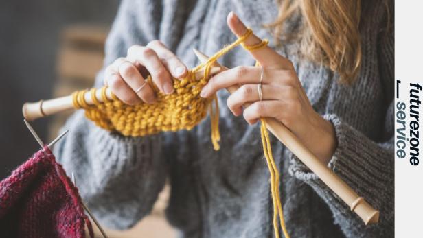 Hände stricken mit dicker gelber Wolle und Holzstricknadeln, grauer Pullover im Hintergrund.