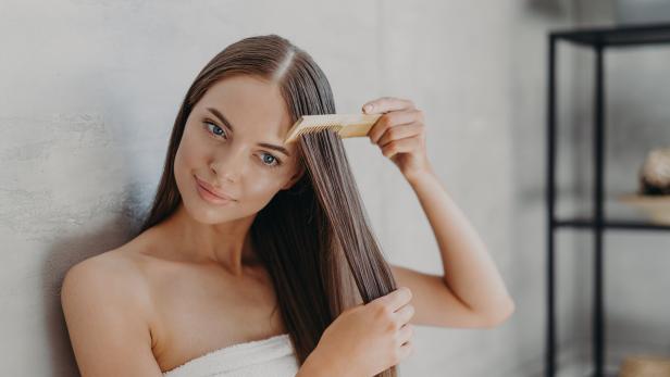 Eine junge Frau mit einem Handtuch um den Körper kämmt ihr langes, braunes Haar.