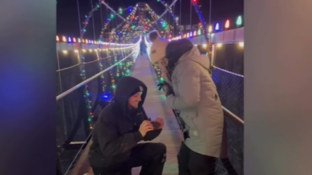 Mann kniet auf beleuchteter Hängebrücke und hält einer Frau einen Ring entgegen.