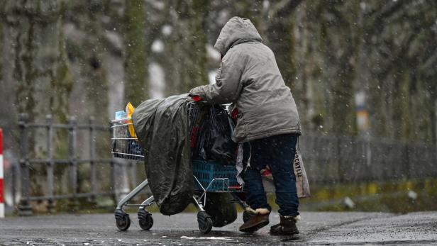 Person in grauer Kapuze schiebt im Schneeregen einen voll beladenen Einkaufswagen.
