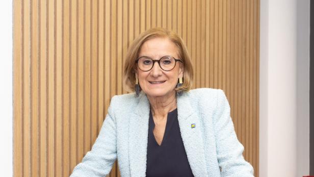 Eine Frau mit Brille und hellblauem Blazer steht lächelnd hinter einer roten Sitzgelegenheit vor einer Holzvertäfelung.