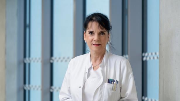 Eine Frau im weißen Arztkittel steht mit Stiften in der Brusttasche vor großen Fenstern und blickt freundlich nach vorn.