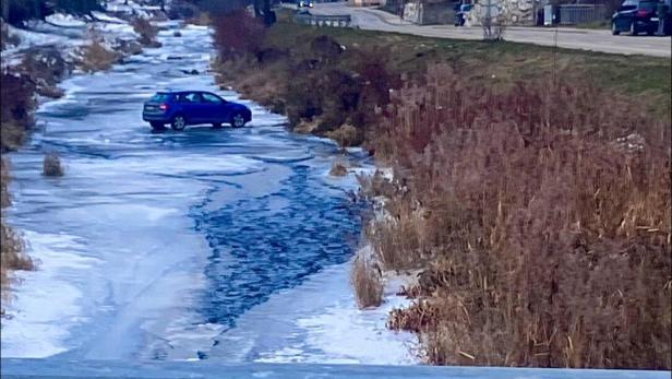 Ein blaues Auto steht mitten auf einem zugefrorenen Fluss, umgeben von winterlicher Landschaft und Häusern am Ufer.