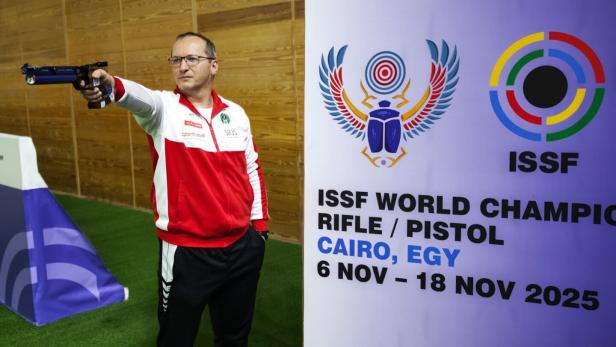 Ein Mann im roten Trainingsanzug zielt mit einer Pistole neben einem Plakat zur ISSF Weltmeisterschaft 2025 in Kairo.