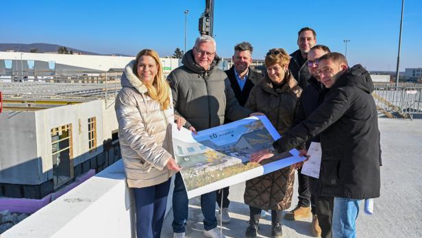 Sieben Personen stehen auf einer Baustelle und betrachten gemeinsam einen großen Architekturplan.