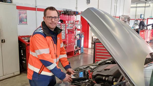 Ein Mann in Warnkleidung prüft mit einem Diagnosegerät den Motor eines Autos in einer Werkstatt.