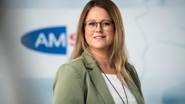 Frau mit langen blonden Haaren, Brille und grünem Blazer steht vor einem unscharfen Hintergrund mit blauem Logo.