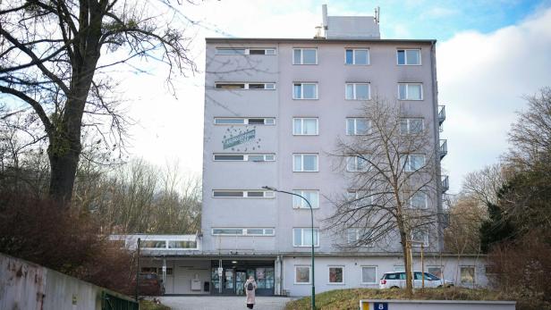 Ein mehrstöckiges graues Gebäude mit vielen Fenstern, davor eine Person auf einem gepflasterten Weg und kahle Bäume.