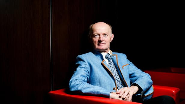 Ein Mann mit blauem Sakko und Krawatte sitzt in einem roten Sessel vor dunklem Hintergrund.