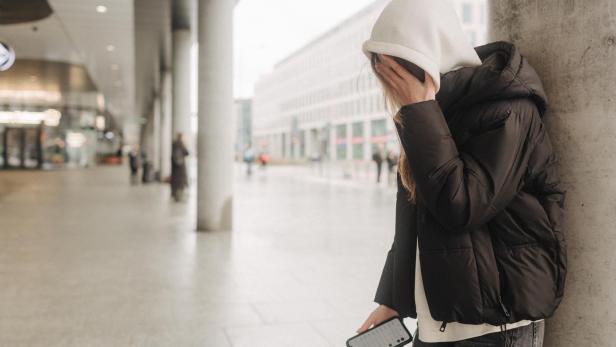 Ein Mädchen mit Kapuze über dem Kopf lehnt an einer Säule, das Smartphone und den Kopf hält.