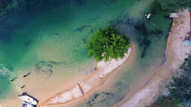 Sandbank mit einem einzelnen Baum im klaren Wasser, Boote am Ufer und Menschen beim Schwimmen und Entspannen.