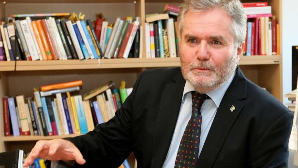 Ein Mann mit Anzug und Krawatte sitzt vor einem Bücherregal und gestikuliert.