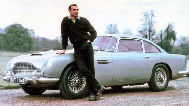 Sean Connery lehnt lässig an einem silbernen Aston Martin DB5 mit dem Kennzeichen BMT 216A.
