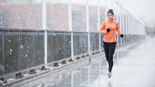 Eine Frau joggt bei Schneefall auf einer Brücke und trägt eine orange Jacke sowie schwarze Leggings.
