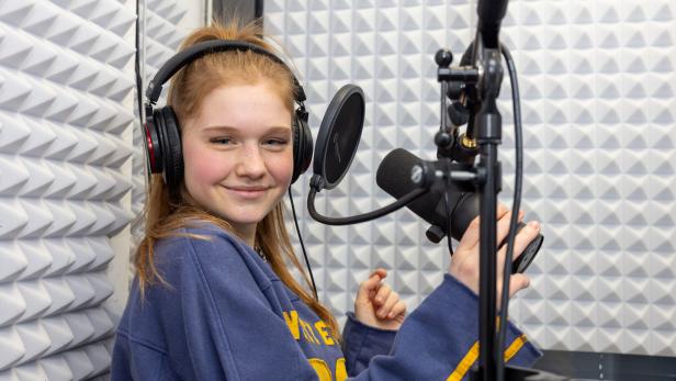 Eine junge Frau mit Kopfhörern lächelt in einem Tonstudio vor einem Mikrofon.