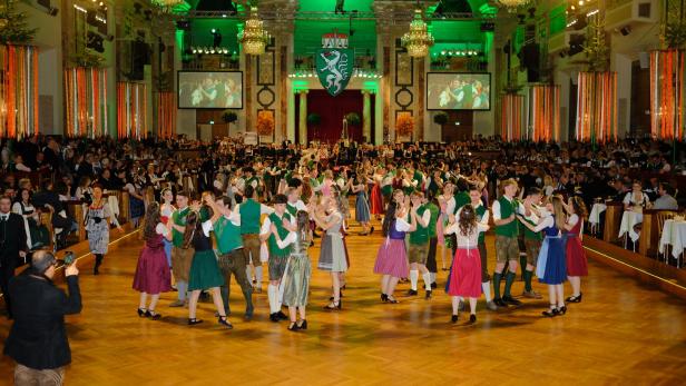 Viele Paare in Tracht tanzen in einem festlich geschmückten Saal mit großem Publikum und Kronleuchtern.