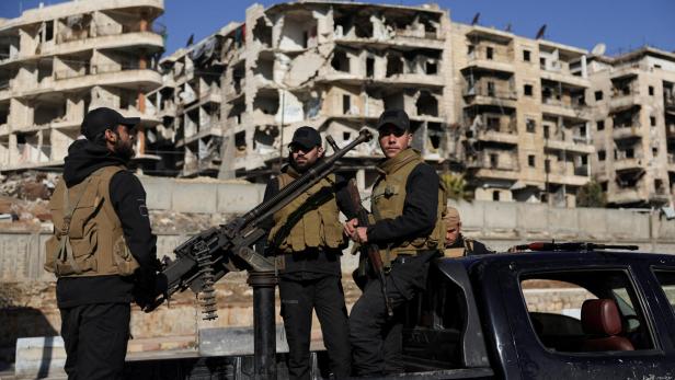 Bewaffnete Männer stehen auf einem Pickup vor zerstörten Gebäuden.