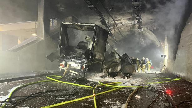 In einem Tunnel steht ein ausgebrannter LKW, umgeben von Löschschaum und Feuerwehrleuten.