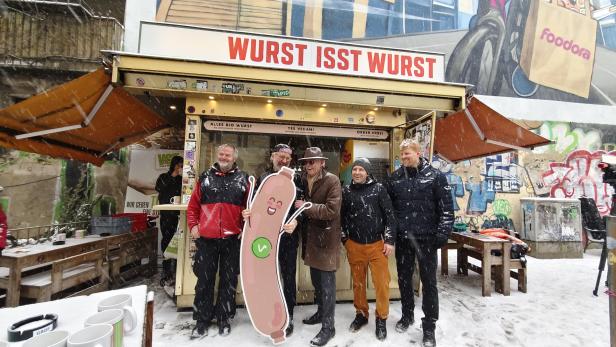 Mehrere Menschen stehen vor dem Würstelstand mit Aufschrift "Wurst isst Wurst"