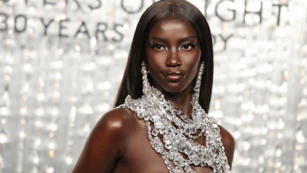 Eine Frau mit glatter, dunkler Haut trägt auffälligen, funkelnden Schmuck und ein silbernes Outfit vor glitzerndem Hintergrund.