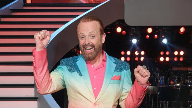 Ein Mann mit Bart trägt einen auffälligen Anzug in Pink und Türkis und posiert fröhlich vor einer beleuchteten Showtreppe.