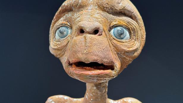 Nahaufnahme von E.T. mit großen, blauen Augen und brauner Haut vor dunklem Hintergrund.