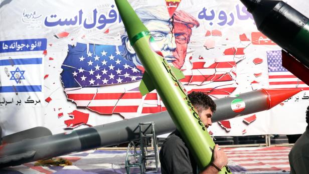 Ein Mann trägt eine grüne Rakete vor einem Banner mit zerrissenen Flaggen der USA und Israels.