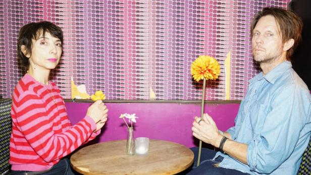 Zwei Personen (Ruth Brauer-Kvam und Kyrre Kvam) sitzen sich an einem kleinen runden Tisch gegenüber und halten jeweils eine gelbe Blume in der Hand.