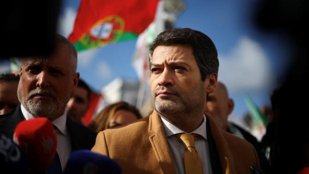 Ein Mann im Anzug steht im Freien vor Mikrofonen, im Hintergrund weht eine portugiesische Flagge.