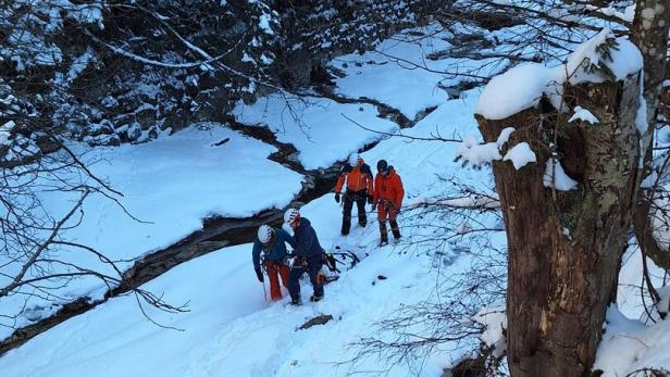 Eine Gruppe von Bergrettern überquert einen schneebedeckten Bach in einer bewaldeten Umgebung.