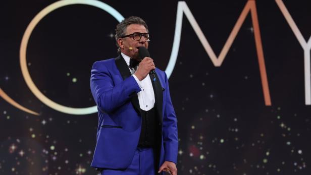 Ein Mann im blauen Anzug spricht in ein Mikrofon vor einem dunklen Hintergrund mit Lichtern.