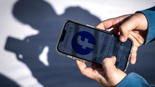 Zwei Hände halten ein Smartphone mit Facebook-Logo auf dem Display, im Hintergrund ist ein Schatten zu sehen.