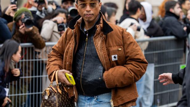 Pharrell Williams trägt eine braune Jacke, Schlaghosen und eine Louis Vuitton Tasche.