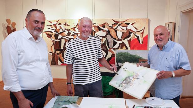 Drei Männer stehen in einer Kunstausstellung, einer hält ein Aquarellbild, auf dem Tisch liegen Malutensilien.