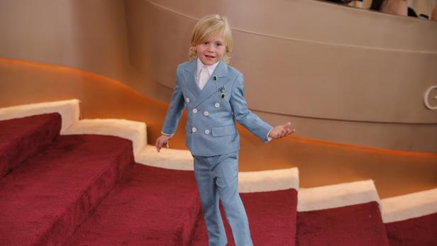 Ein blondes Kind im hellblauen Anzug steht auf einer roten Treppe und lächelt mit ausgestrecktem Arm.
