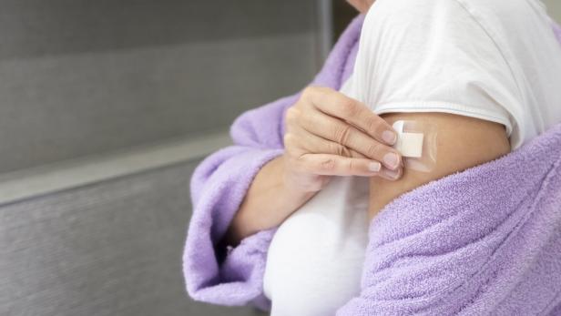 Eine Person mit weißem T-Shirt und lila Bademantel klebt ein Pflaster auf ihren Oberarm.