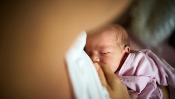 Ein Neugeborenes in rosa Kleidung wird gestillt und liegt eng an eine erwachsene Person geschmiegt.
