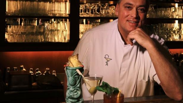 Barchef Heinz Kaiser lächelt hinter seiner Bar mit drei seiner Mocktails und vollen Spirituosenregalen.