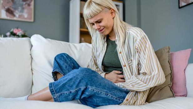 Eine junge Frau sitzt auf einem Sofa und hält sich mit schmerzverzerrtem Gesicht den Bauch.