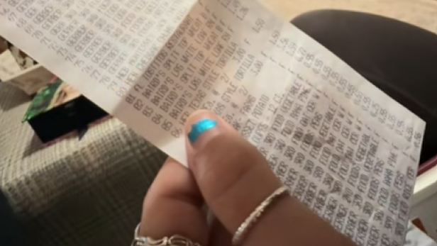 Hand hält einen langen, verblassten Kassenzettel in einem Wohnzimmer mit Fernseher im Hintergrund.