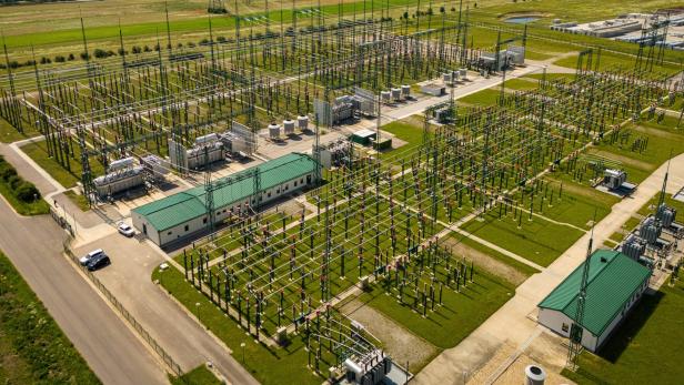 Große Umspannanlage mit zahlreichen Stromleitungen, Transformatoren und Gebäuden auf grünem Gelände.