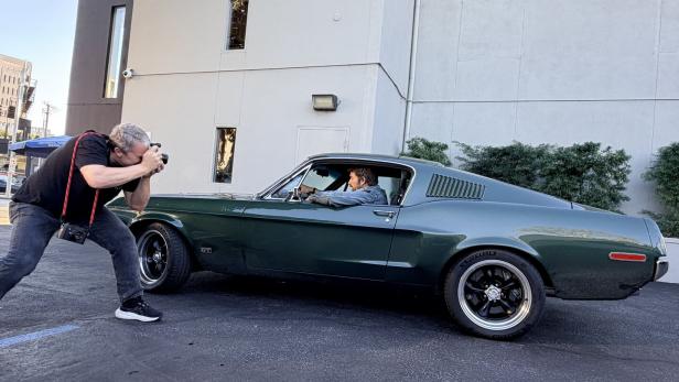 Ein Fotograf macht ein Bild von einer Person, die in einem dunkelgrünen Oldtimer vor einem Gebäude sitzt.