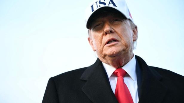 Donald Trump mit "USA"-Kappe, dunklem Anzug und roter Krawatte vor hellem Hintergrund.