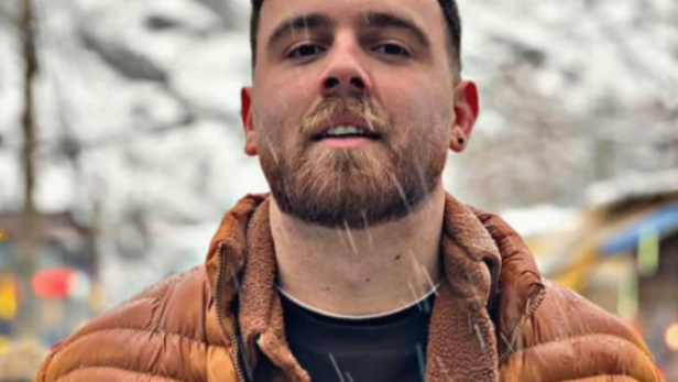 Ein junger Mann mit Bart steht im Schnee und trägt eine braune Winterjacke.