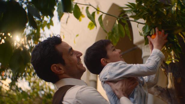 Ein Mann hebt einen Buben in die Höhe, damit er eine Orange pflücken kann.