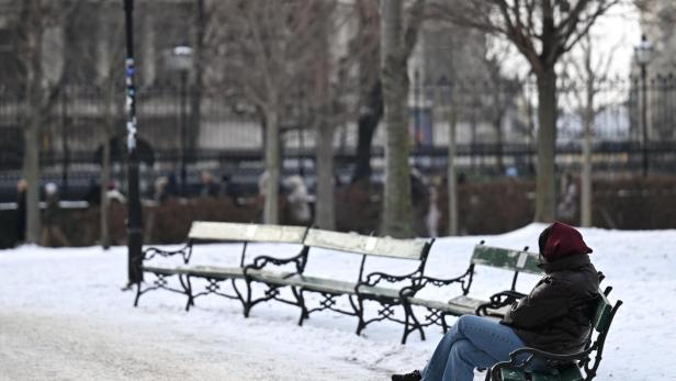 Eine Person sitzt allein auf einer Parkbank im verschneiten Park, umgeben von kahlen Bäumen und leeren Bänken.