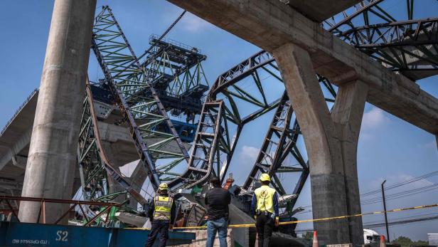 Drei Personen stehen vor einer eingestürzten Brückenkonstruktion aus Stahl und Beton, umgeben von Absperrband.