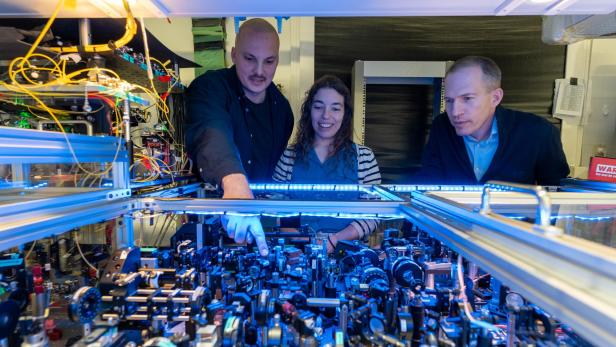 Drei Personen stehen an einem komplexen optischen Experimentaufbau mit zahlreichen Linsen, Spiegeln und Kabeln in einem Labor.