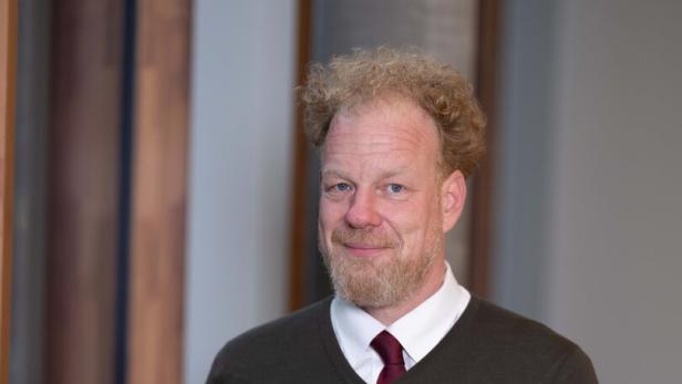 Ein Mann mit lockigem, blondem Haar und Bart trägt einen dunkelgrünen Pullover über einem Hemd mit Krawatte.